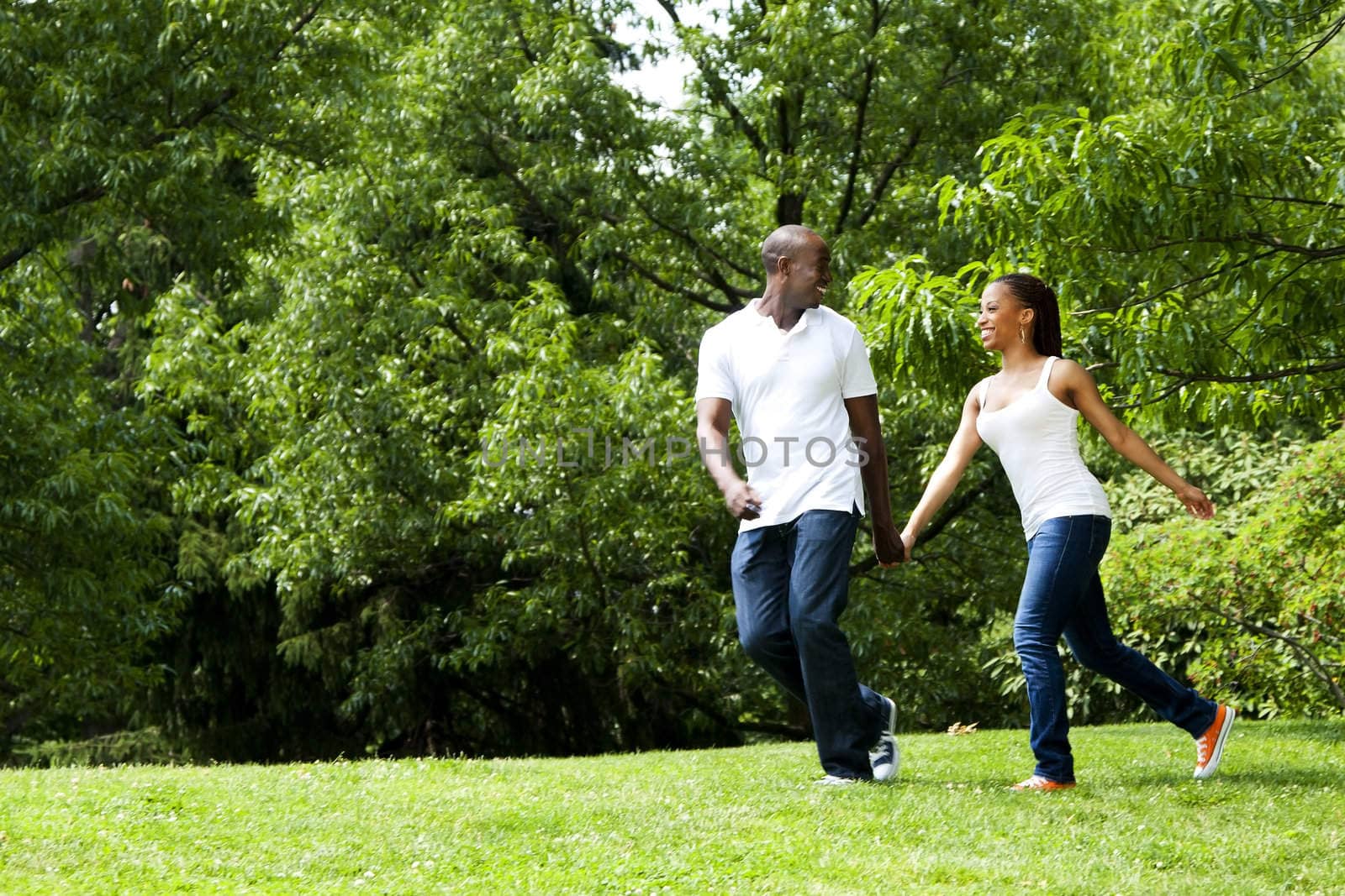 Beautiful fun happy smiling African American young couple running playing in park, wearing white shirts and blue jeans.