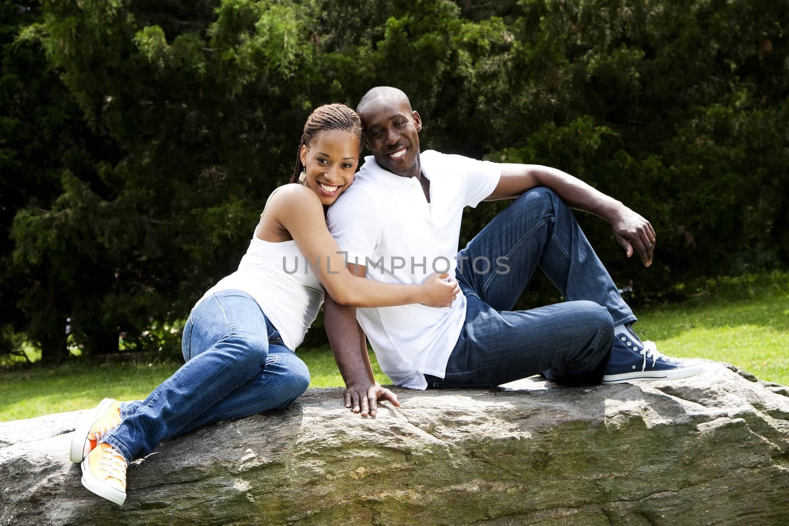 Beautiful fun happy smiling African American couple in love wearing white shirts and blue jeans, sitting on a rock in a park, woman hugging guy.
