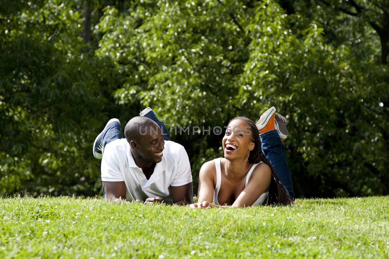 Beautiful fun happy smiling laughing African American couple joking laying on grass in park, wearing white shirts and blue jeans.