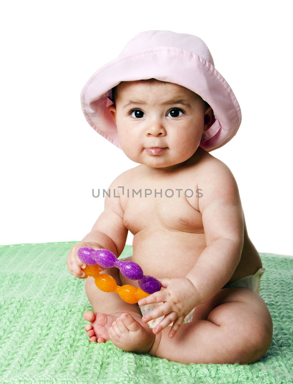 Beautiful cute Caucasian Hispanic baby girl sitting on green blanket holding an orange with purple teething ring toy, wearing pink hat, isolated.