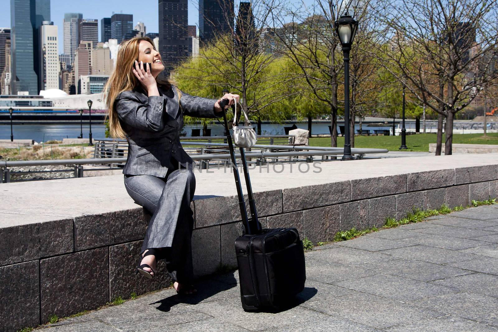 Beautiful happy laughing Caucasian Latina entrepreneur business woman in grey suit sitting in park in New York City talking on cell phone with a carry-on luggage and purse.