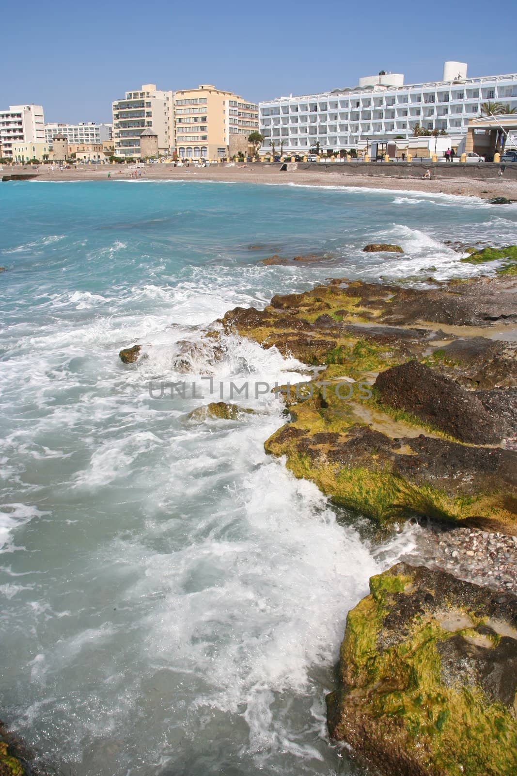 Mediterranean beach with rocks and seaweed, backdrop of hotels on Rhodes coast.