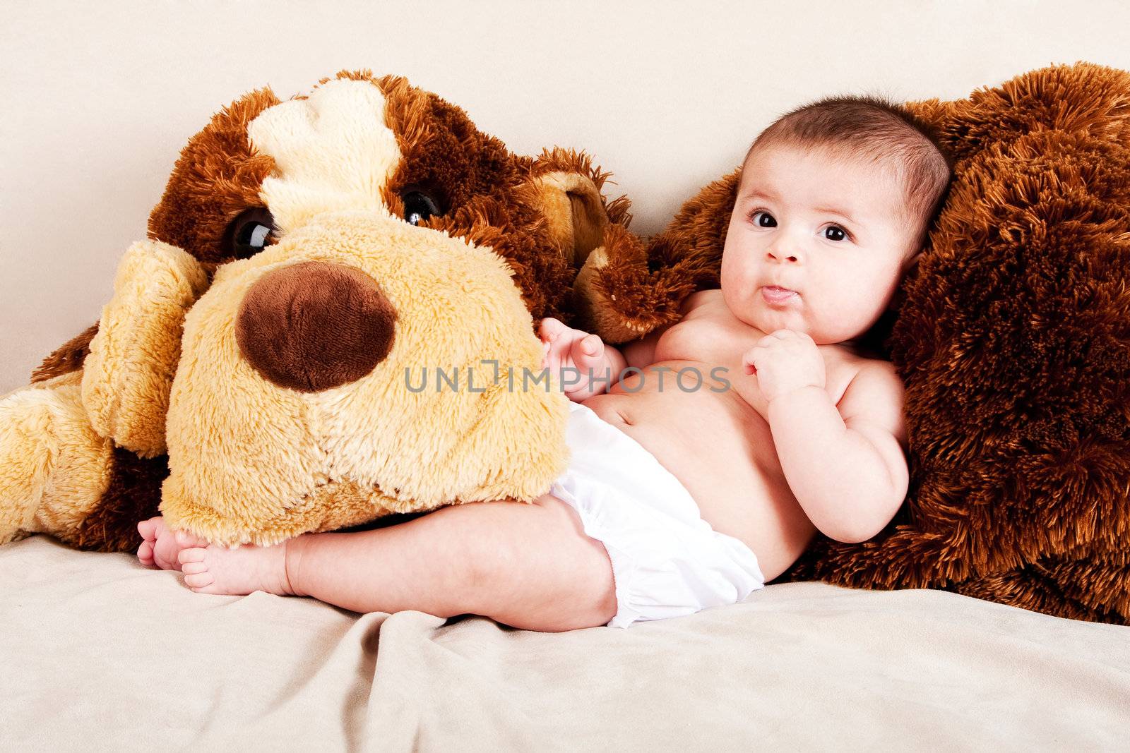 Cute Caucasian Hispanic unisex baby in arms of a big brown stuffed dog laying on couch.