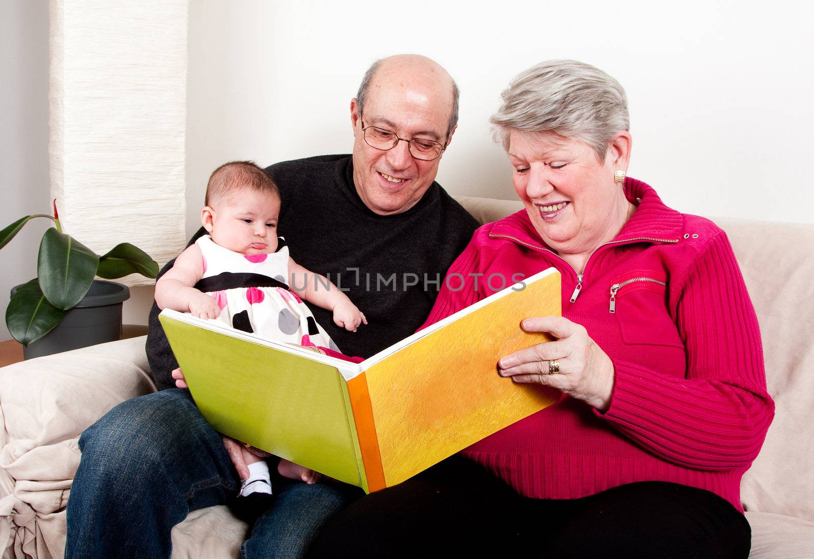 Proud and happy grandparents reading book to educate their grandchild. Baby girl enjoying book while sitting on couch in livingroom.