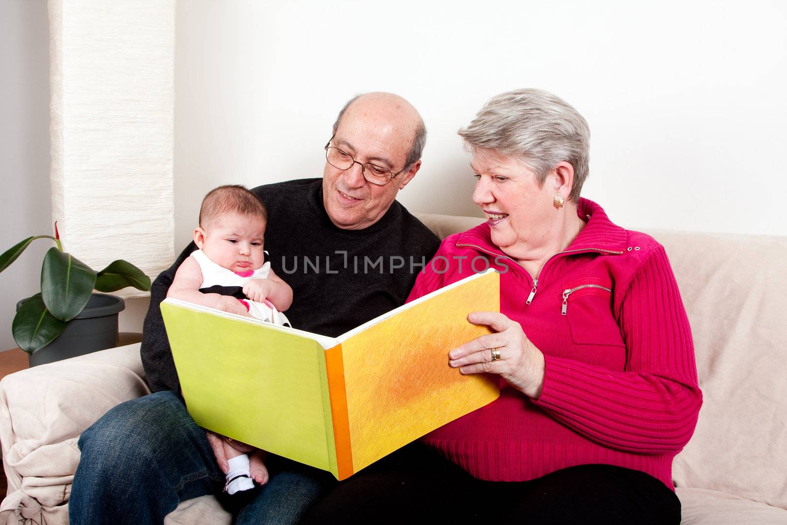 Proud and happy grandparents reading book to educate their grandchild. Baby girl enjoying book while sitting on couch in livingroom.
