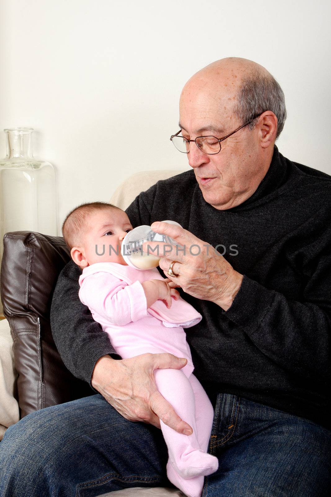 Caucasian Hispanic Grandfather feeding cute baby girl with a bottle of milk while sitting on a couch in a livingroom holding infant in his arms.