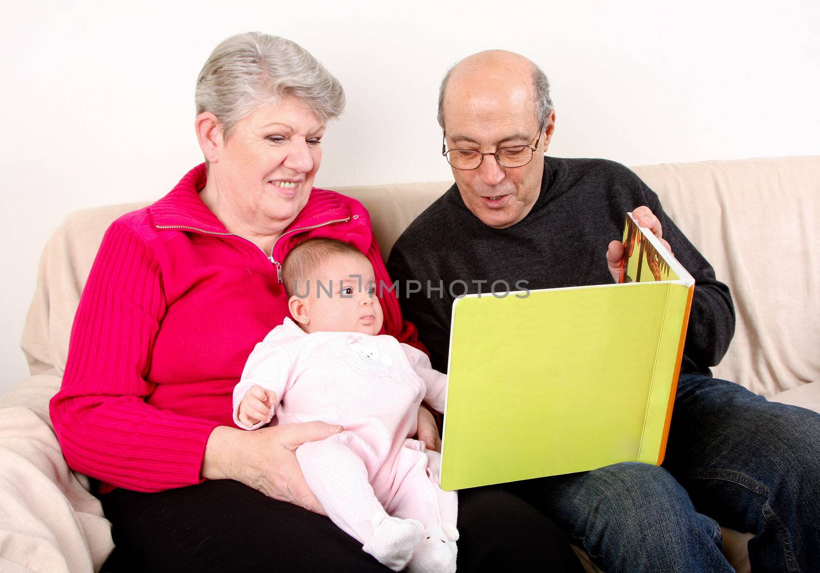 Happy fun Caucasian Hispanic Middle Eastern family sitting together on couch reading book to baby. Grandfather and Grandmother reading green book to infant granddaughter.