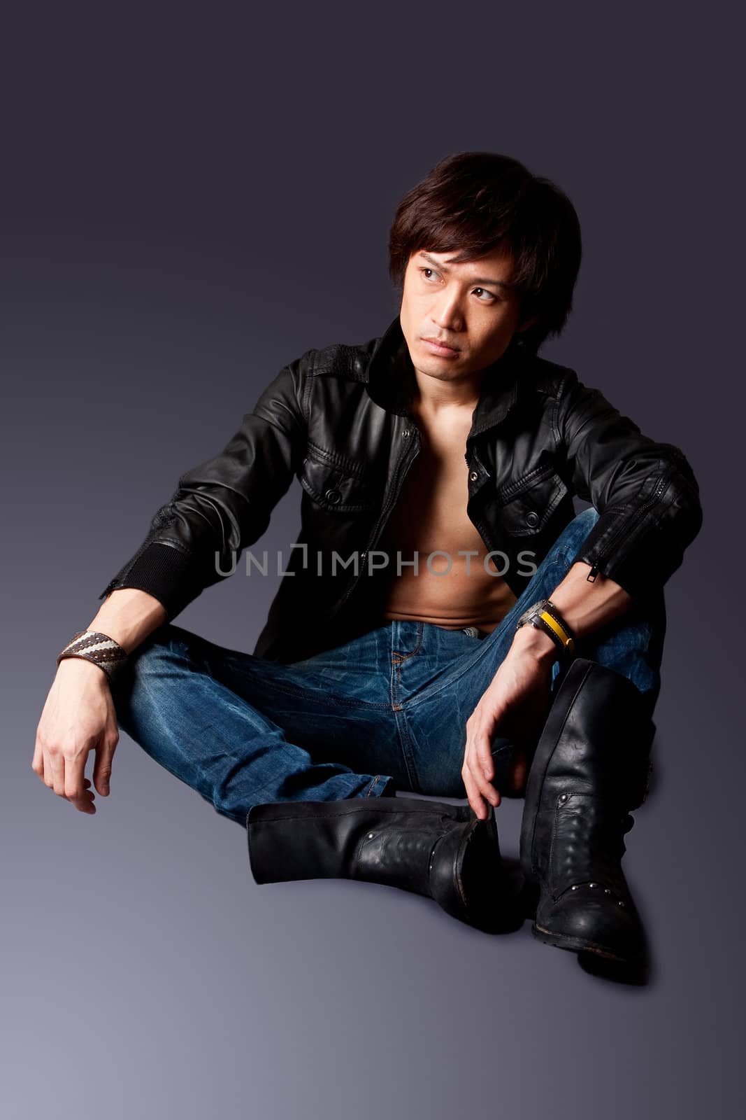 Handsome Asian male wearing leather jacket over a bare chest and jeans with macho attitude while sitting on floor expressing thought, isolated.