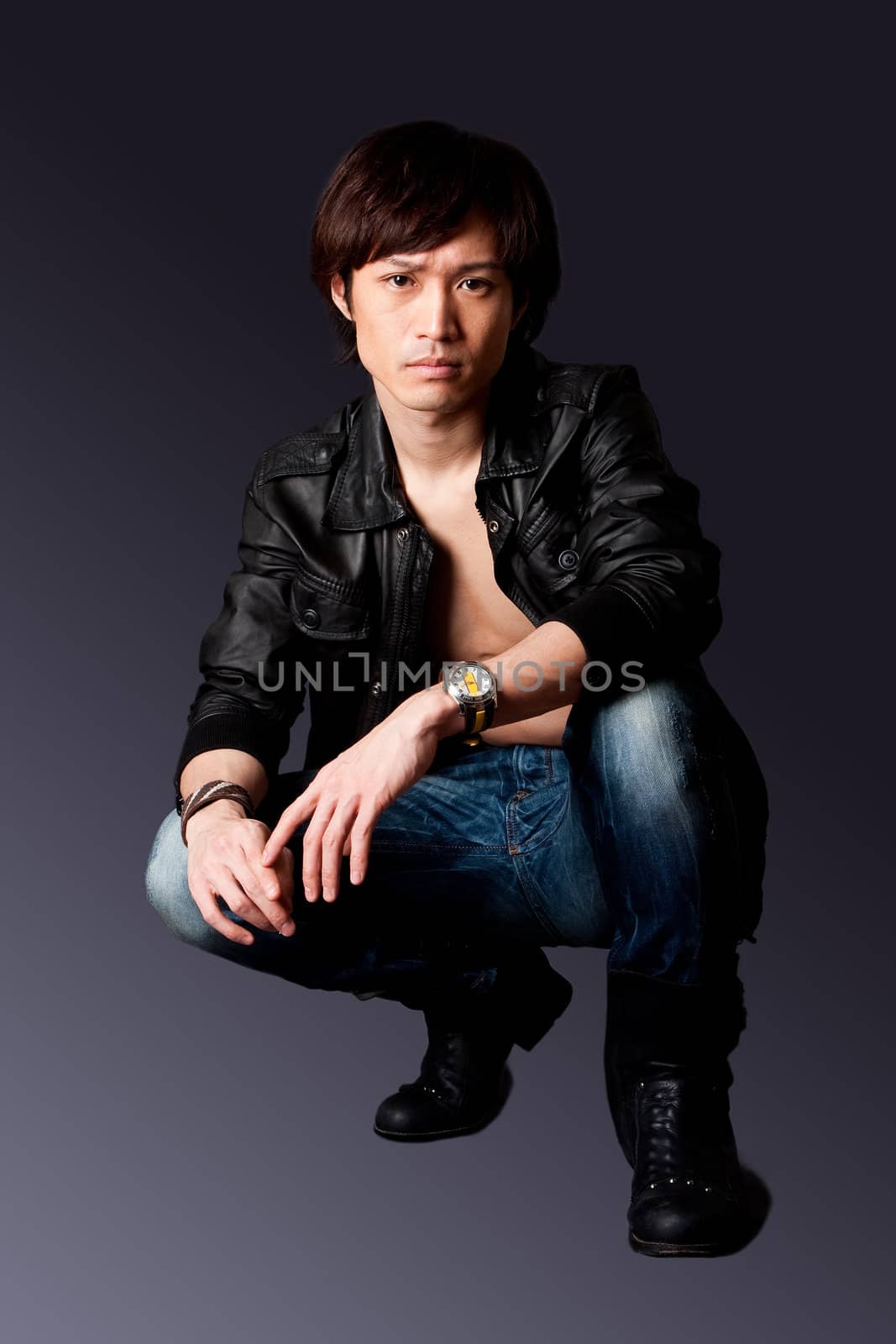 Handsome Asian male wearing leather jacket over a bare chest and jeans with macho attitude while crouching, isolated.