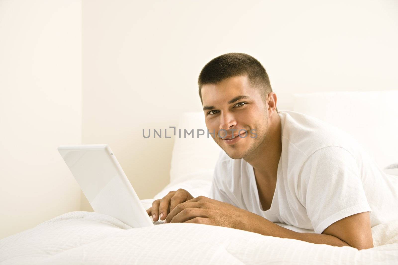 Handsome Caucasian mid adult man lying in bed with laptop looking at viewer.