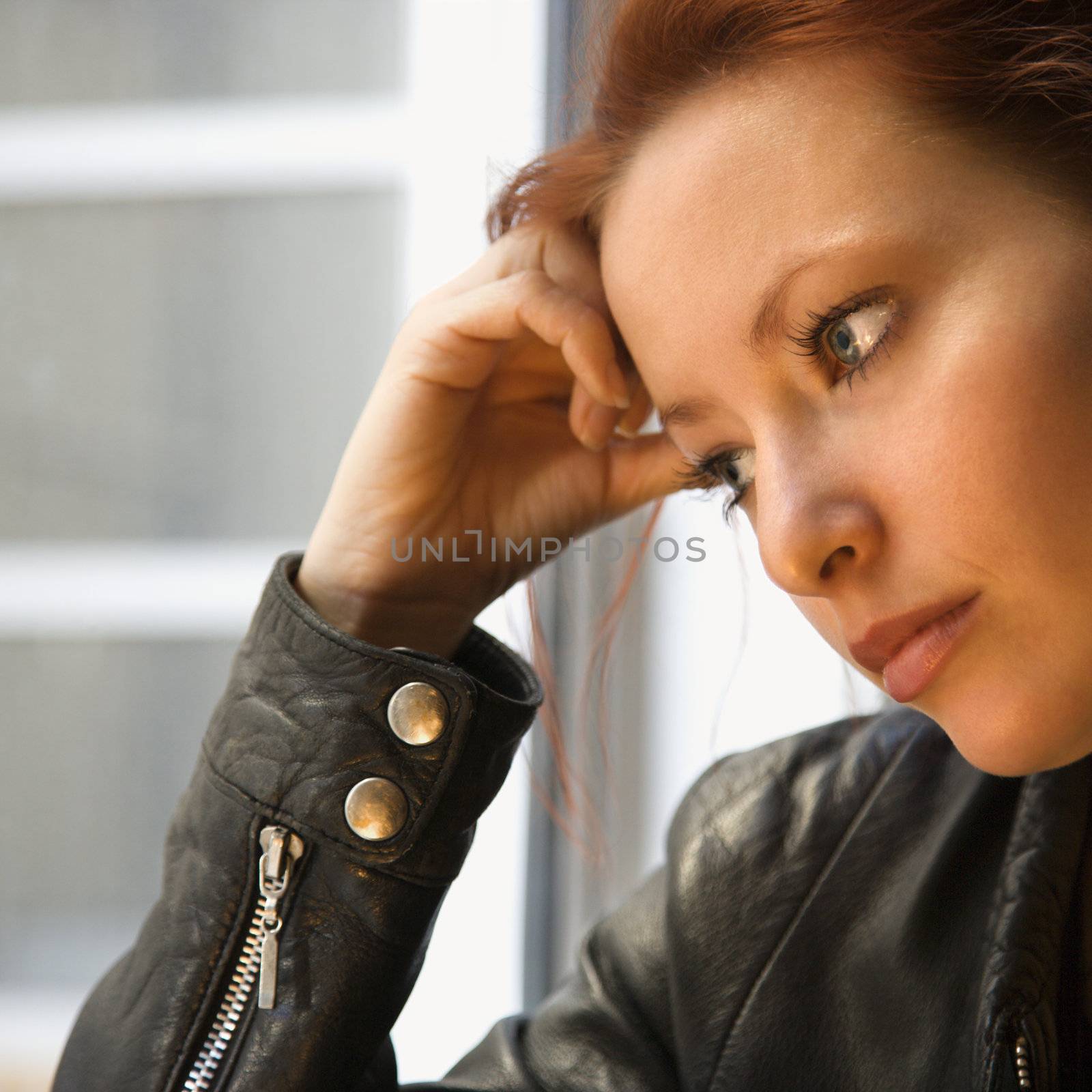 Pretty redhead young woman indoors holding head on hand looking out window.