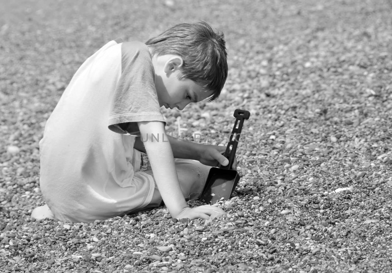 little boy sitting on pebble playing with plastic shovel in black and white