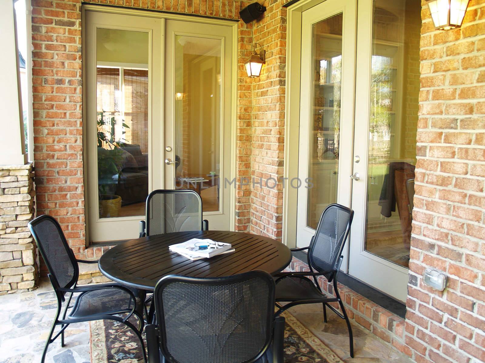 A wood and metal table and chairs on a covered stone patio on an american brick home. Two large glass french doors open up to the house in the background