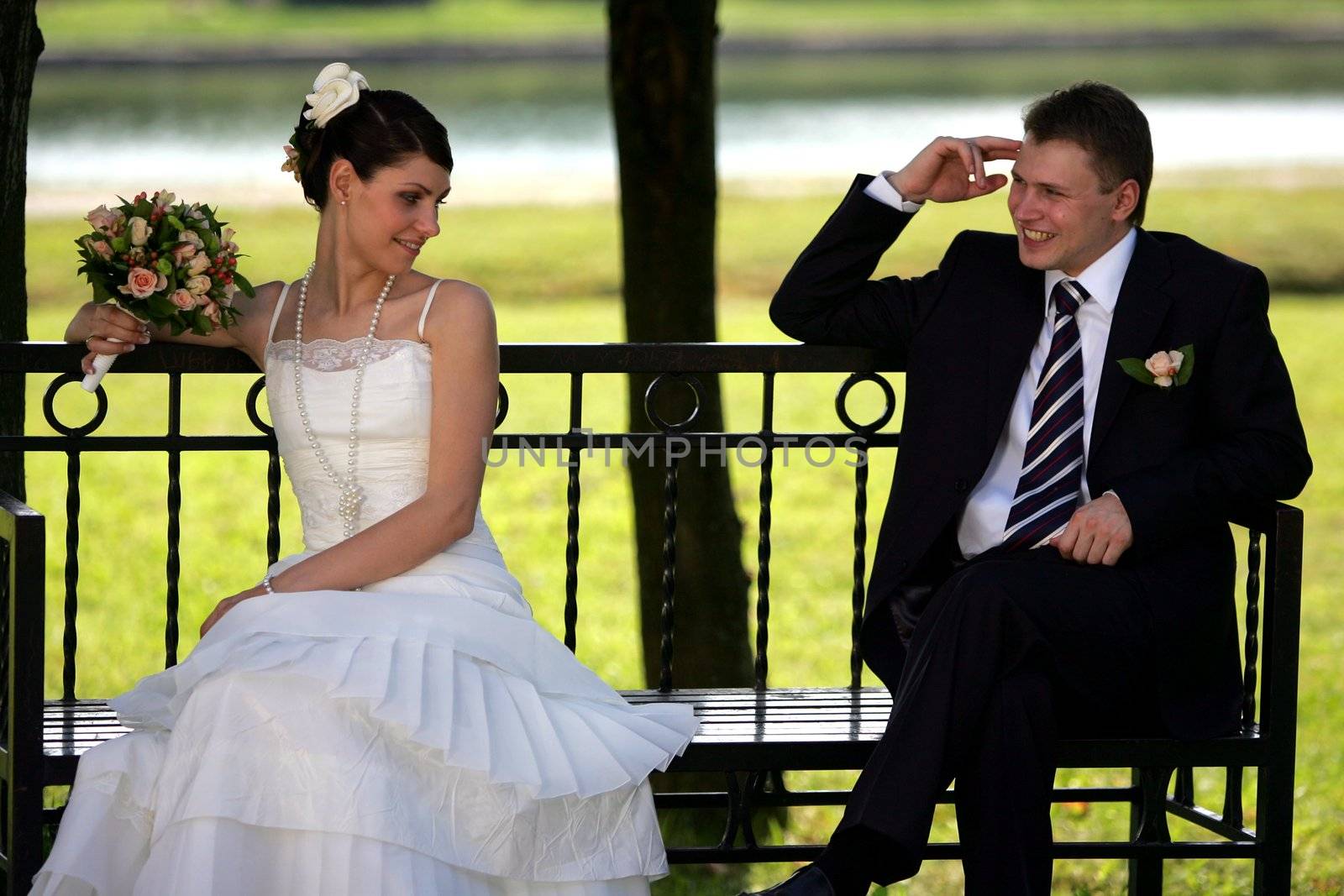 Newlywed couple sat on park bench, smiling groom and coy bride with bouquet.