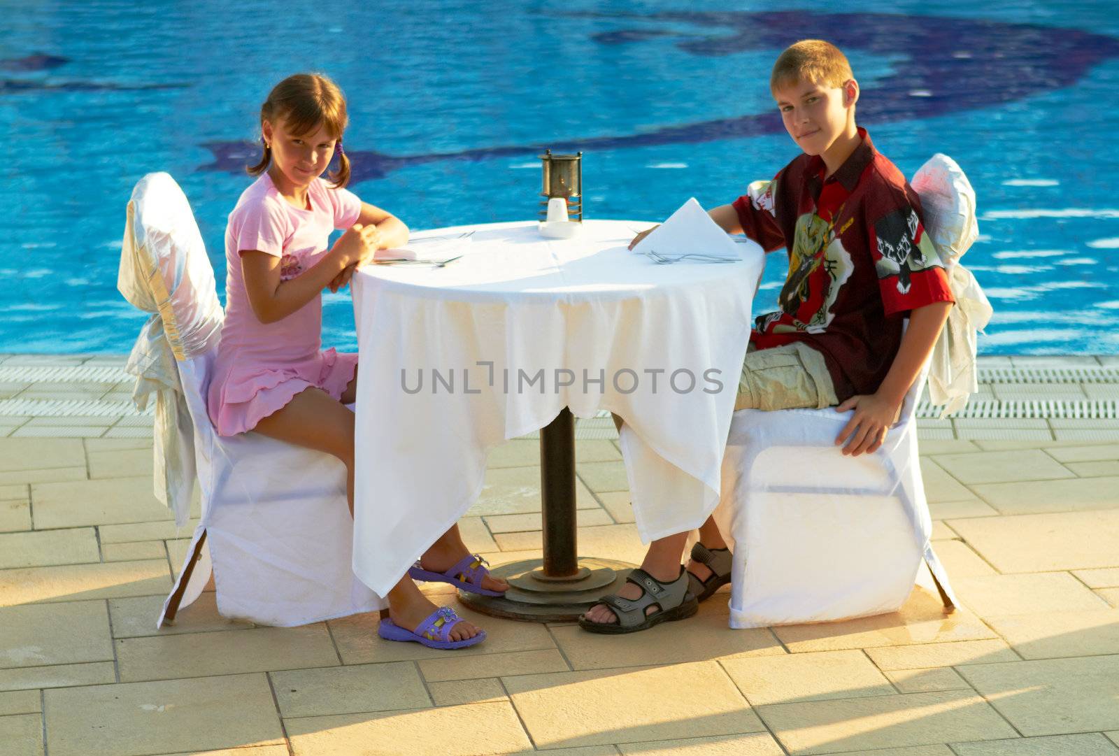 Boy and girl sitting at the restaurant table on the pool side