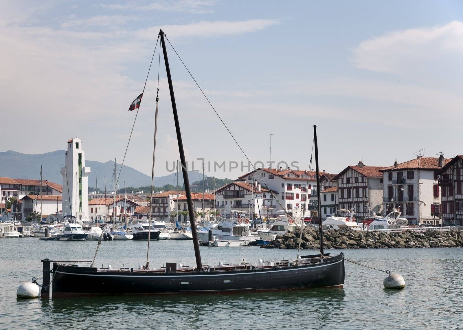 One black boat with one mast and a marina in the background