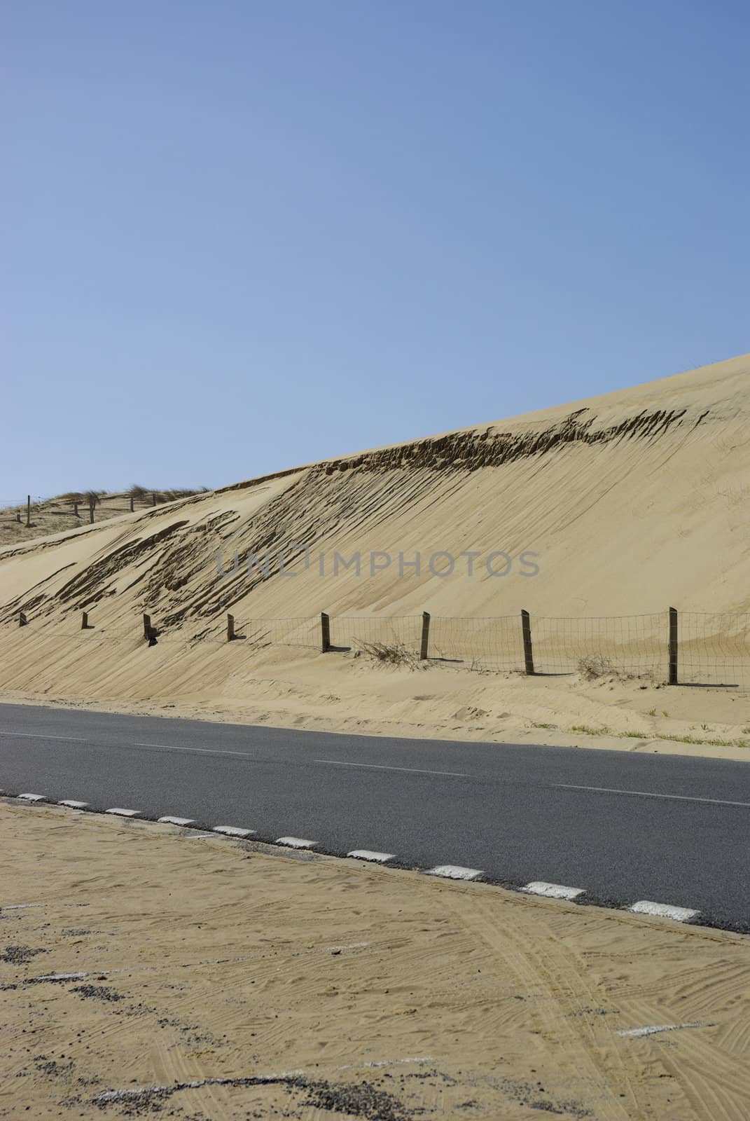 An isolated road along big dunes with a little barrier and some stranding in the sand