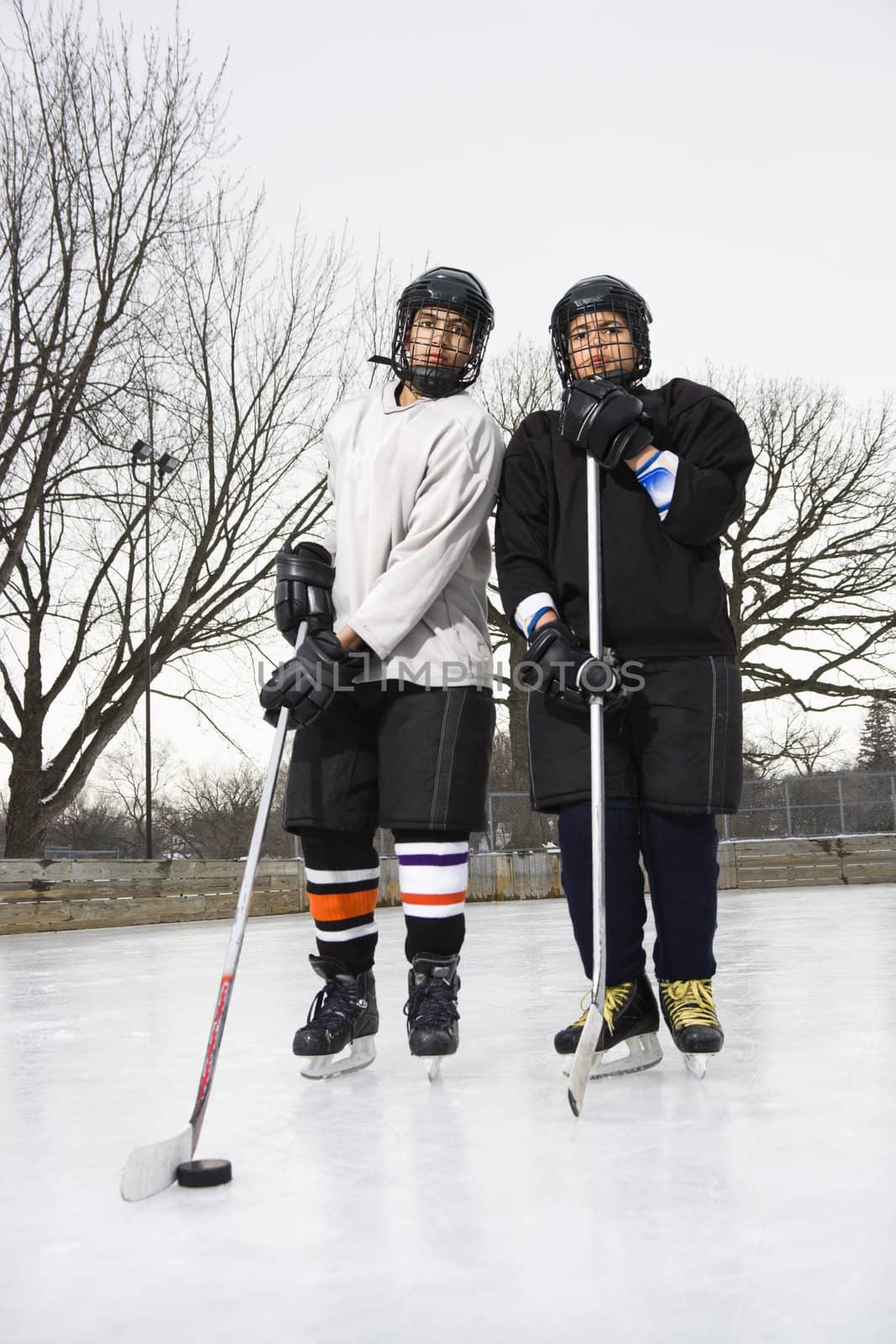 Two boys in ice hockey uniforms holding hockey sticks standing on ice rink in ice skates.