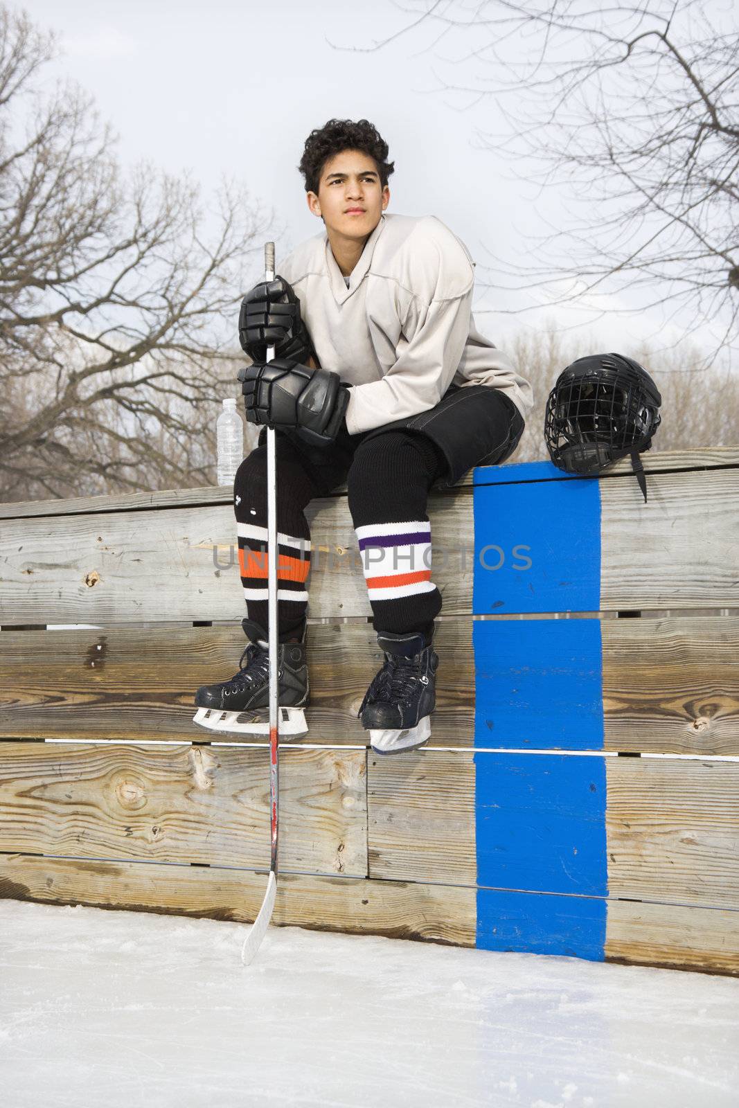 Boy in ice hockey uniform holding hockey stick sitting on sidelines.