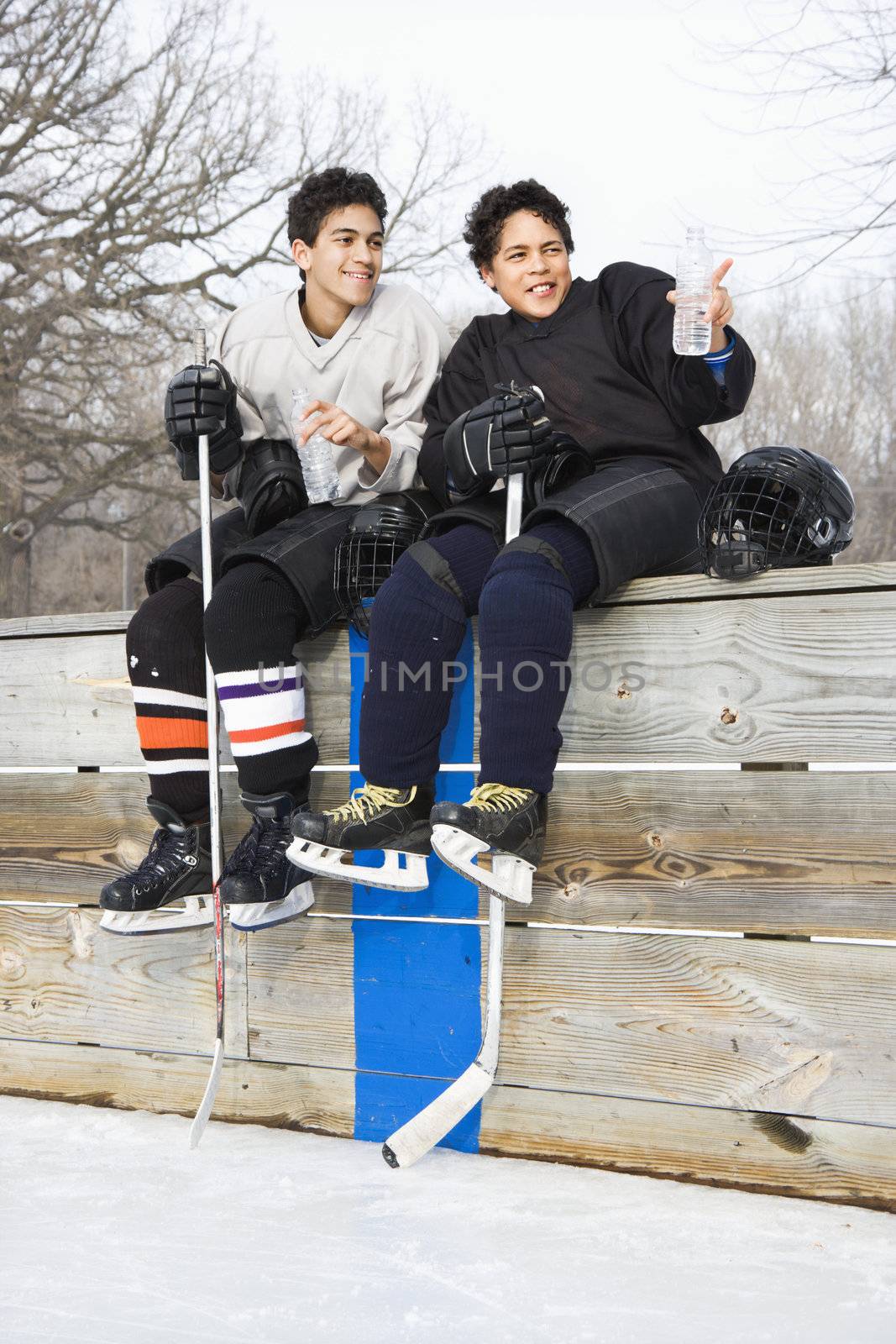 Two boys in ice hockey uniforms sitting on ice rink sidelines pointing and looking.
