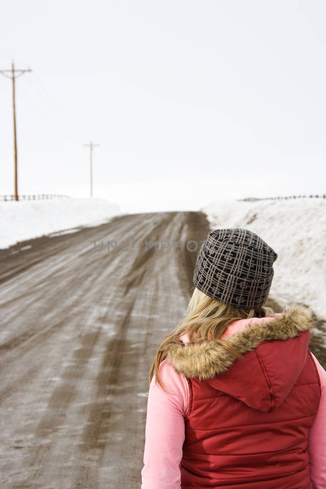 Young woman in winter clothes standing alone looking down a muddy dirt road.