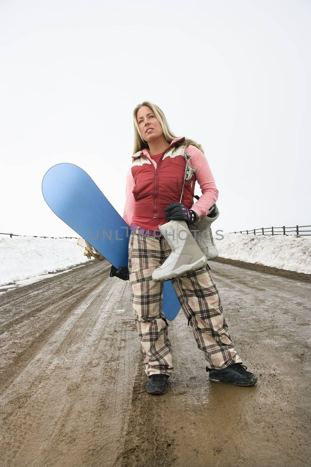 Young woman in winter clothes standing alone on muddy dirt road holding snowboard and boots.