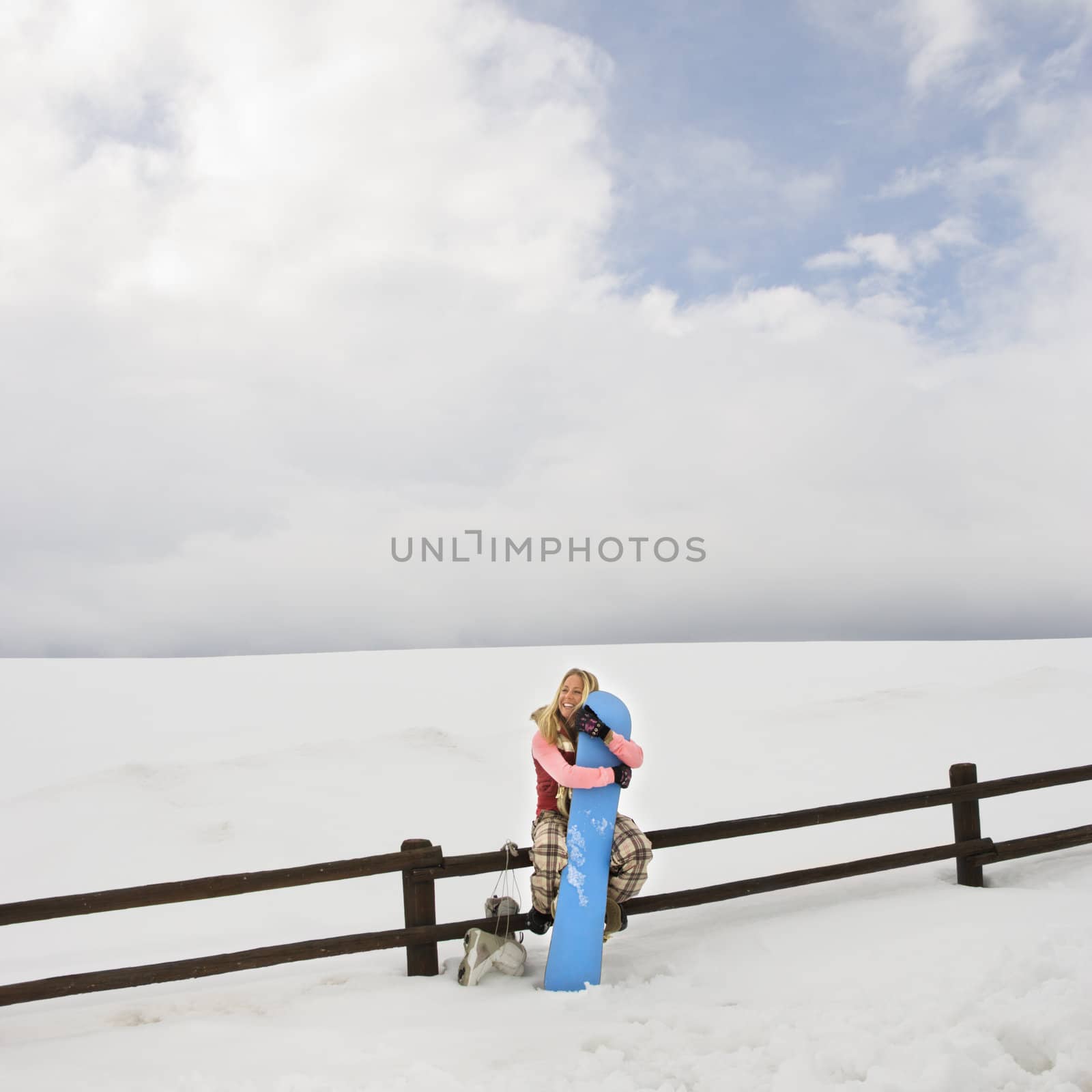 Young woman in winter clothes sitting on fence in snowy field holding snowboard.
