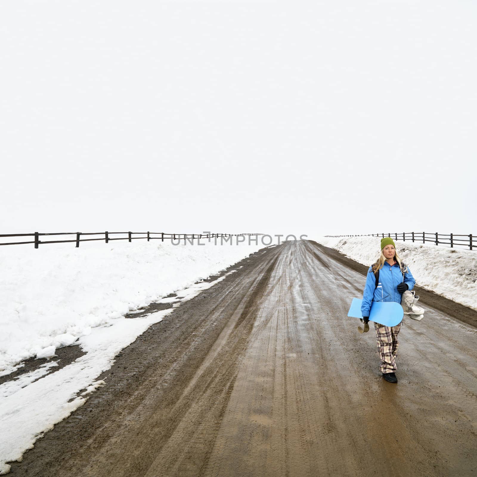 Young woman in winter clothes walking alone down muddy dirt road holding snowboard and boots.