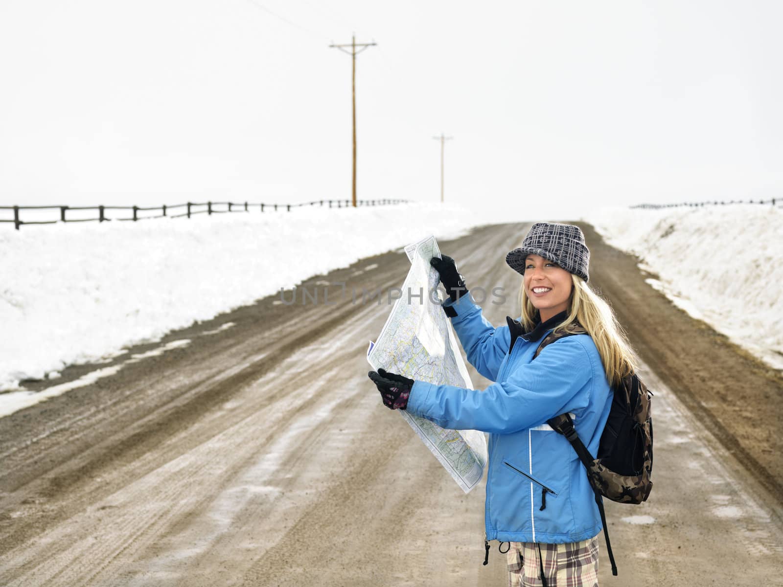 Young woman in winter clothes standing on muddy dirt road holding open map smiling.
