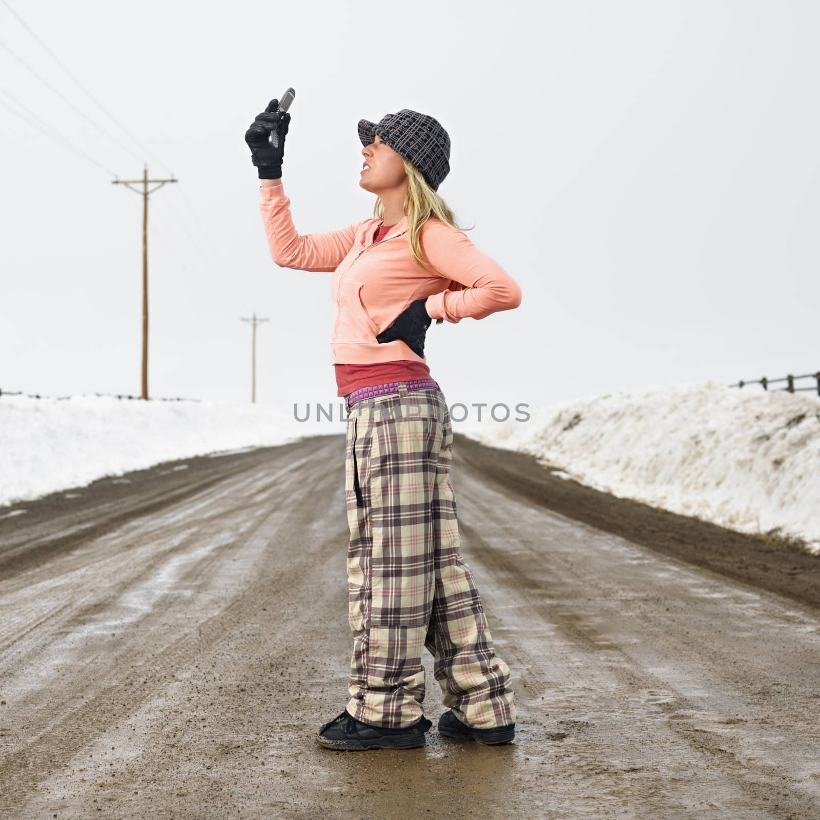 Young woman in winter clothes standing on muddy dirt road looking at cell phone.