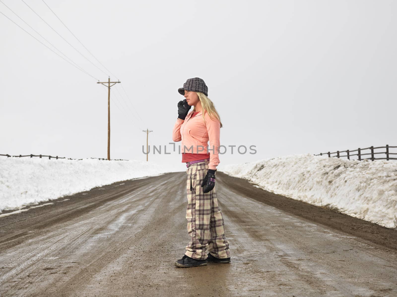 Young woman in winter clothes standing on muddy dirt road talking on cell phone.