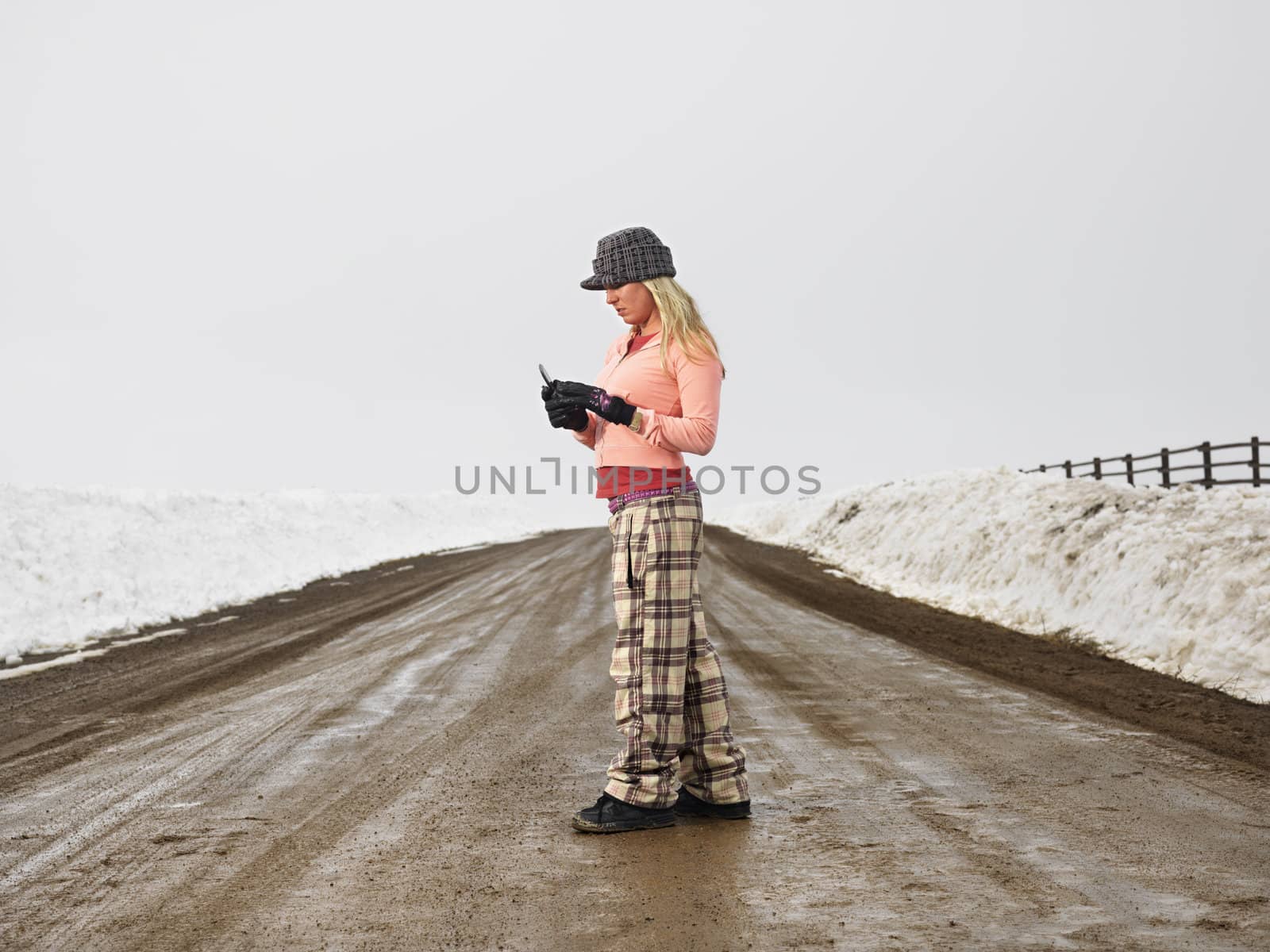 Young woman in winter clothes standing on muddy dirt road looking at cell phone.