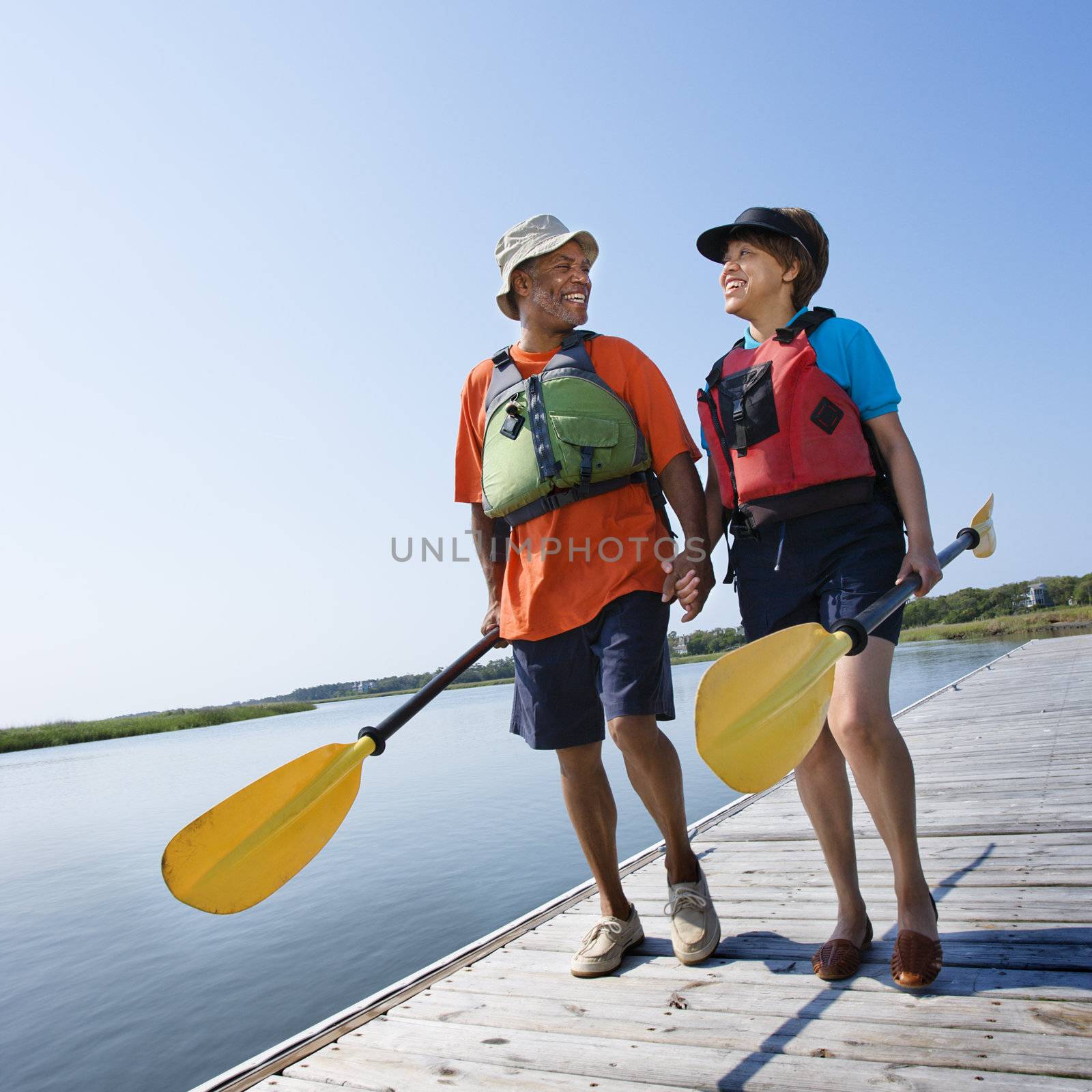 African American middle-aged couple walking on boat dock holding hands and carrying paddles.