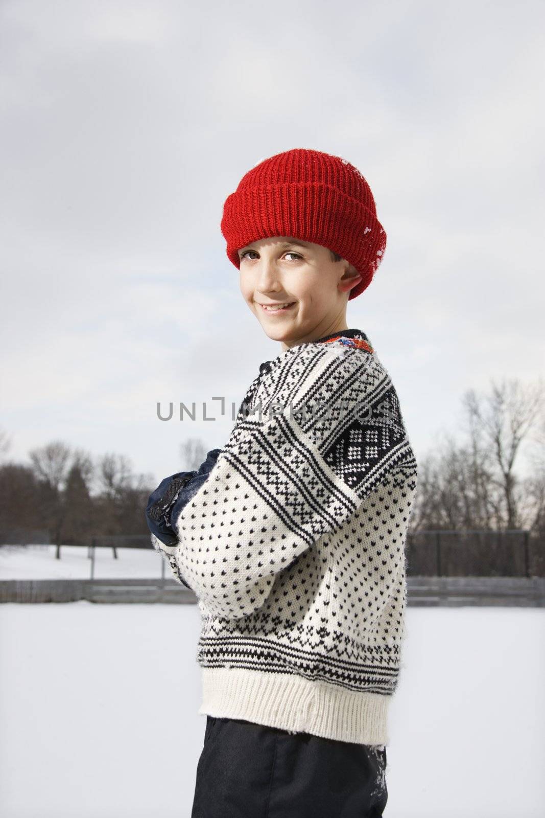 Portrait of Caucasian boy wearing sweater and red winter cap standing looking over shoulder and smiling at viewer.