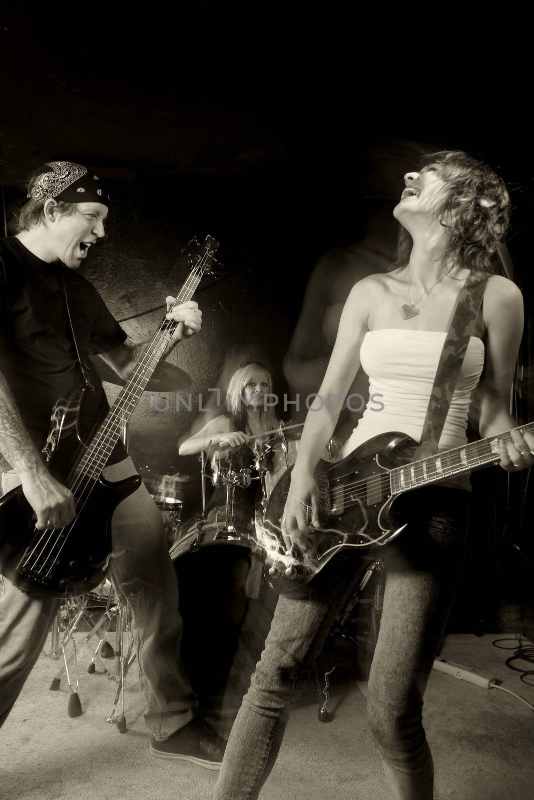 Band playing on a stage. Male bassist with female guitarist and drummer. Shot with strobes and slow shutter speed to create lighting atmosphere and blur effects. Slight motion blur on performers.