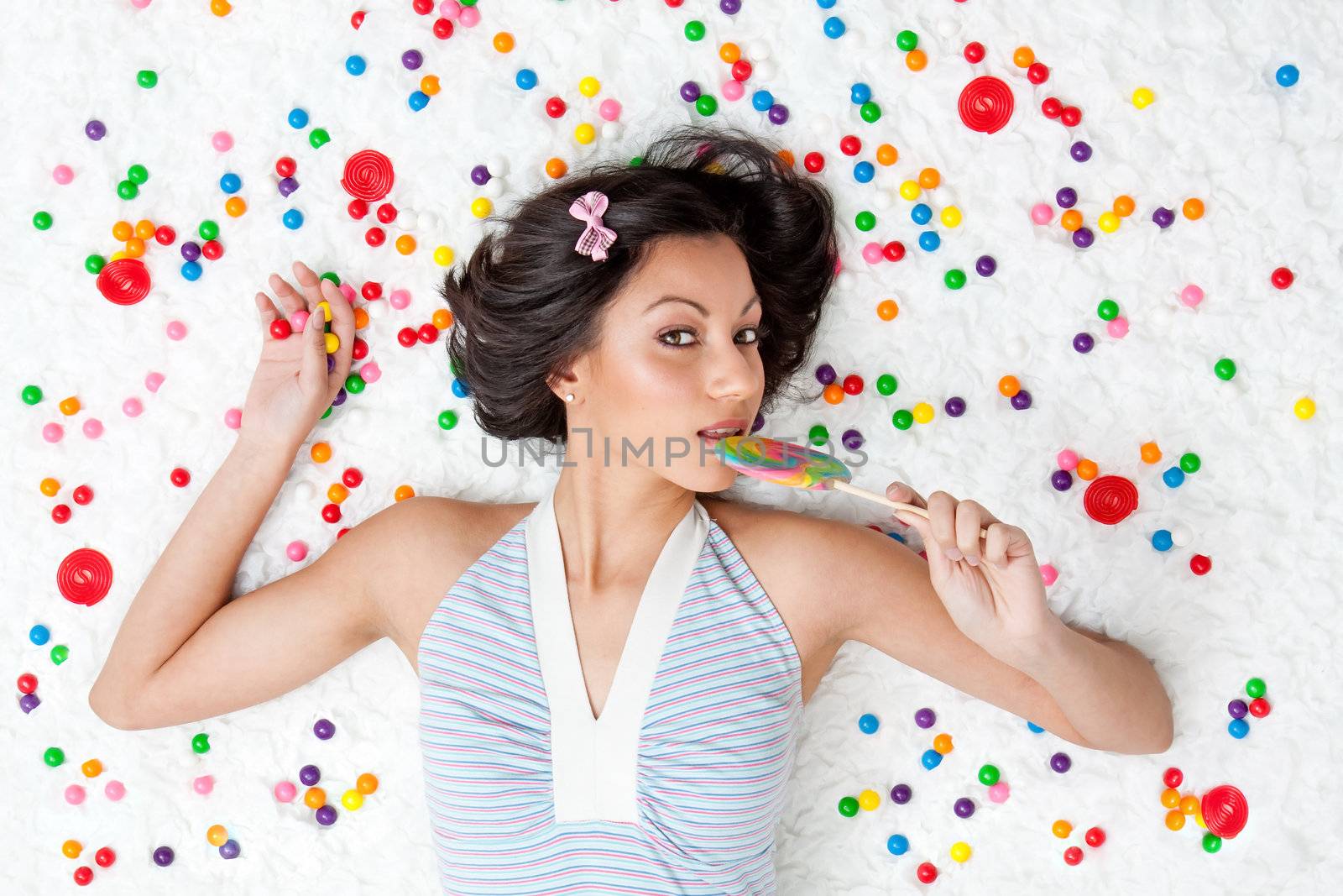 Young Latina woman laying on ruffled cloud like floor between colorful bubblegum balls eating a lollipop