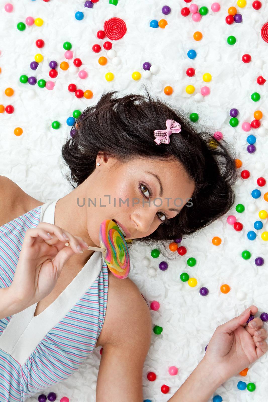 Young Latina woman laying on ruffled cloud like floor between colorful bubblegum balls eating a lollipop