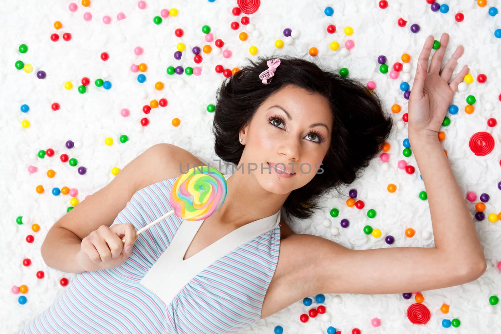 Young Latina woman laying on ruffled cloud like floor between colorful bubblegum balls holding a lollipop