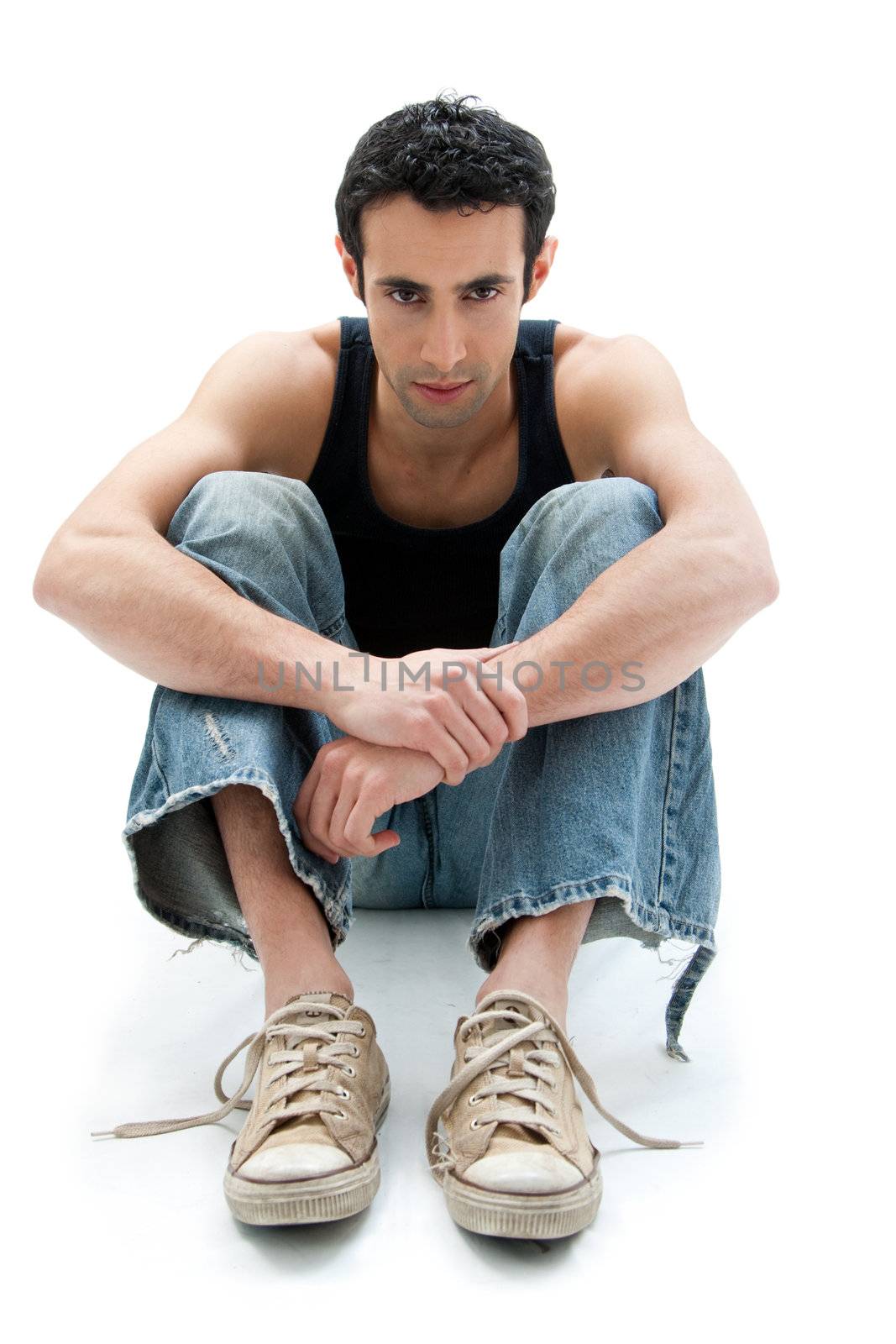 Handsome Caucasian guy wearing black tank top and jeans sitting on floor with arms around knees, isolated
