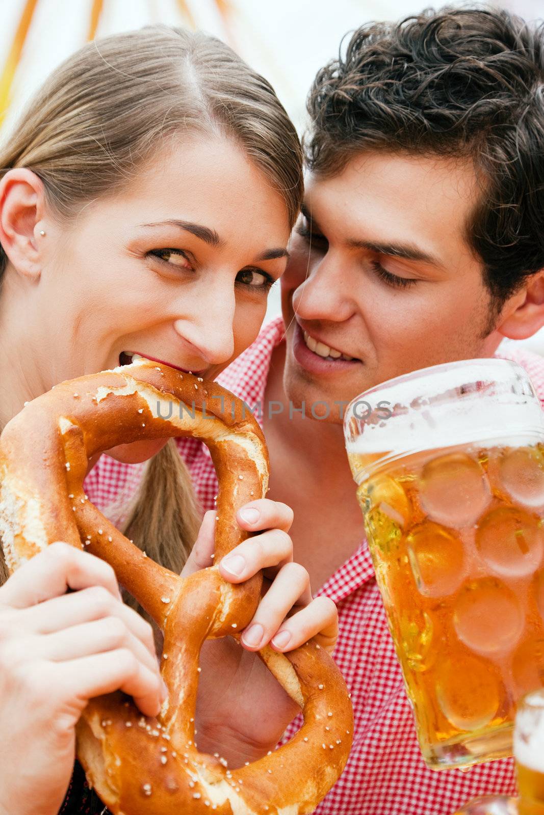 Couple in traditional German costume in a beer tent, he is having a drink, she a pretzel, scene could be located at the Oktoberfest or any Duld