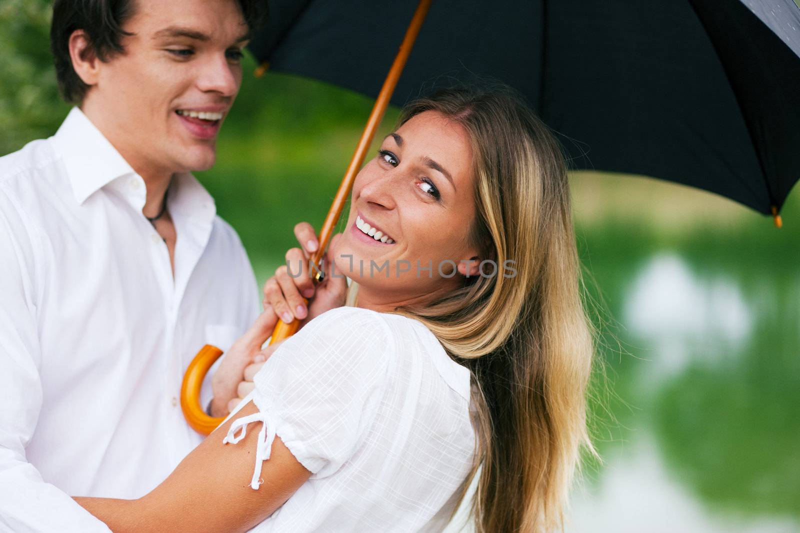 Couple (man and woman) at a lake in the summer rain with an umbrella, laughing and having fun despite of the bad weather