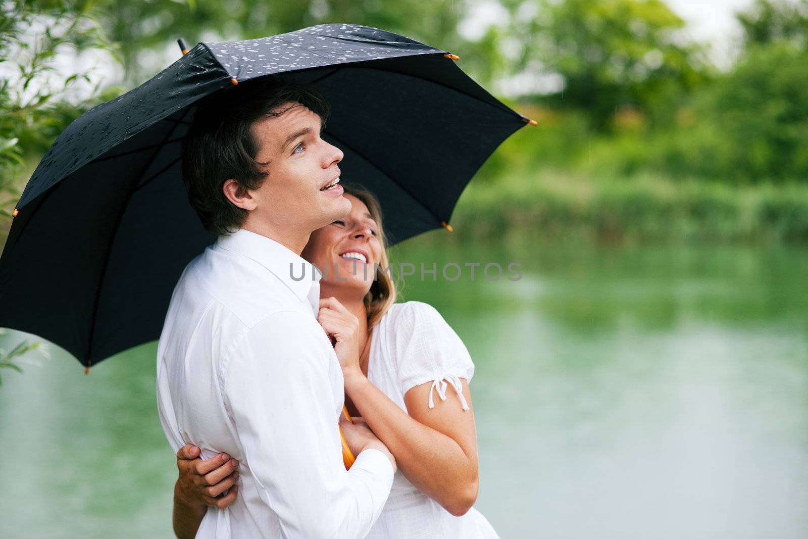 Couple (man and woman) at a lake in summer rain with an umbrella, he is sheltering her from the drops, holding his girl in his arms