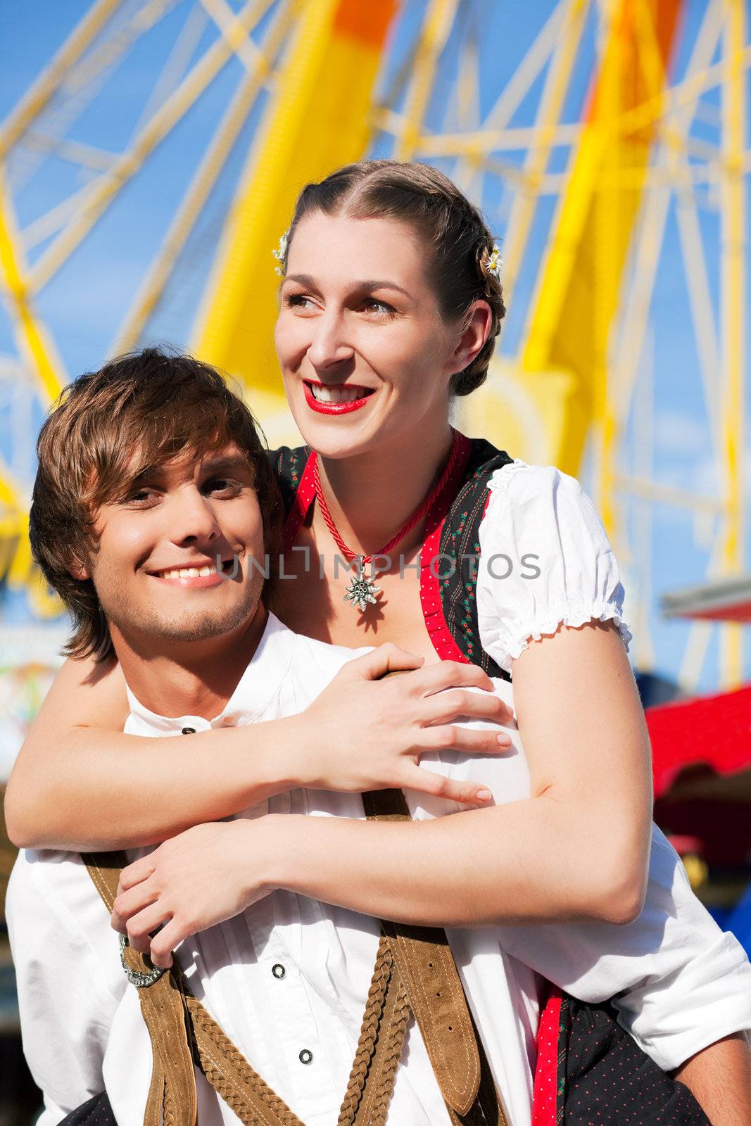 Young and beautiful couple in traditional Bavarian Tracht - Dirndl and Lederhosen - embracing each other on a fair like a Dult or the Oktoberfest; both are standing in front of a typical booth