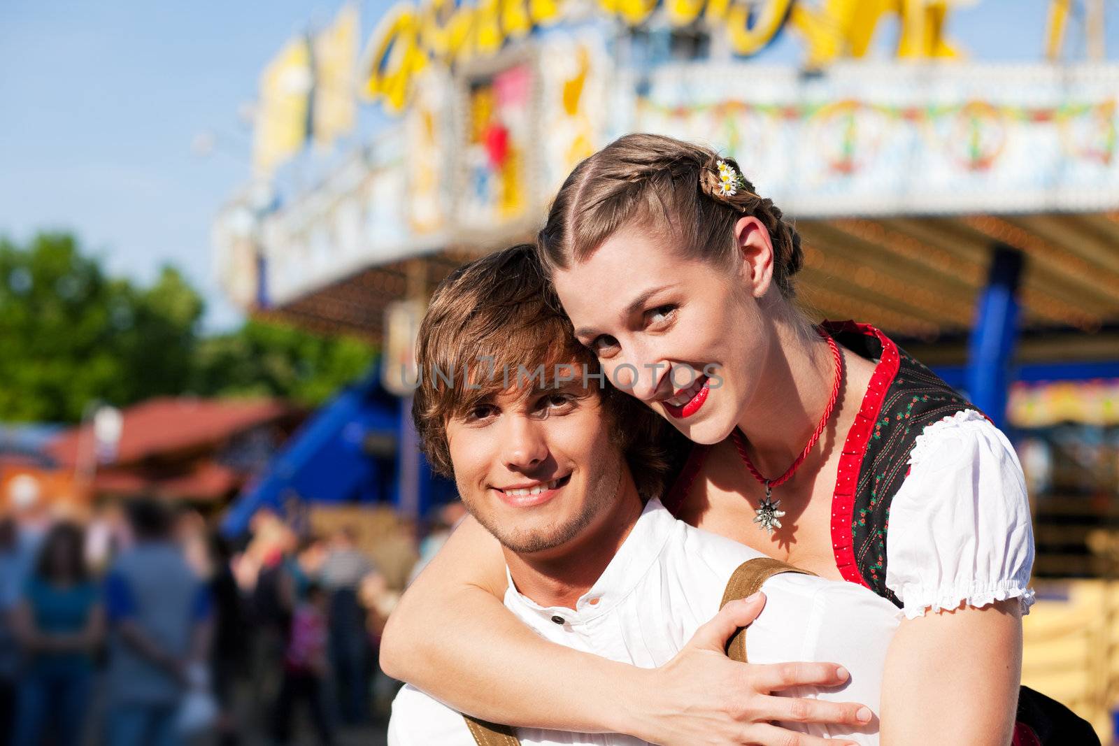 Young and beautiful couple in traditional Bavarian Tracht - Dirndl and Lederhosen - embracing each other on a fair like a Dult or the Oktoberfest; both are standing in front of a big wheel