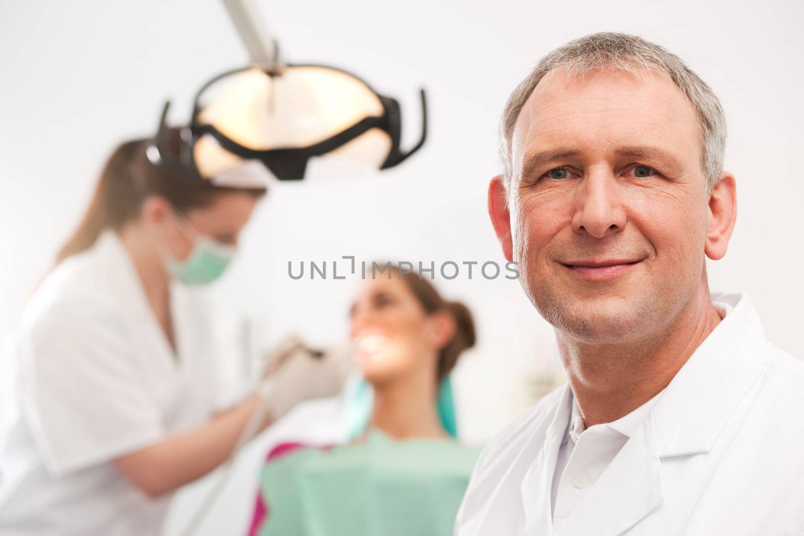 Dentist in his surgery looking at the viewer, in the background his assistant is giving a female patient a treatment