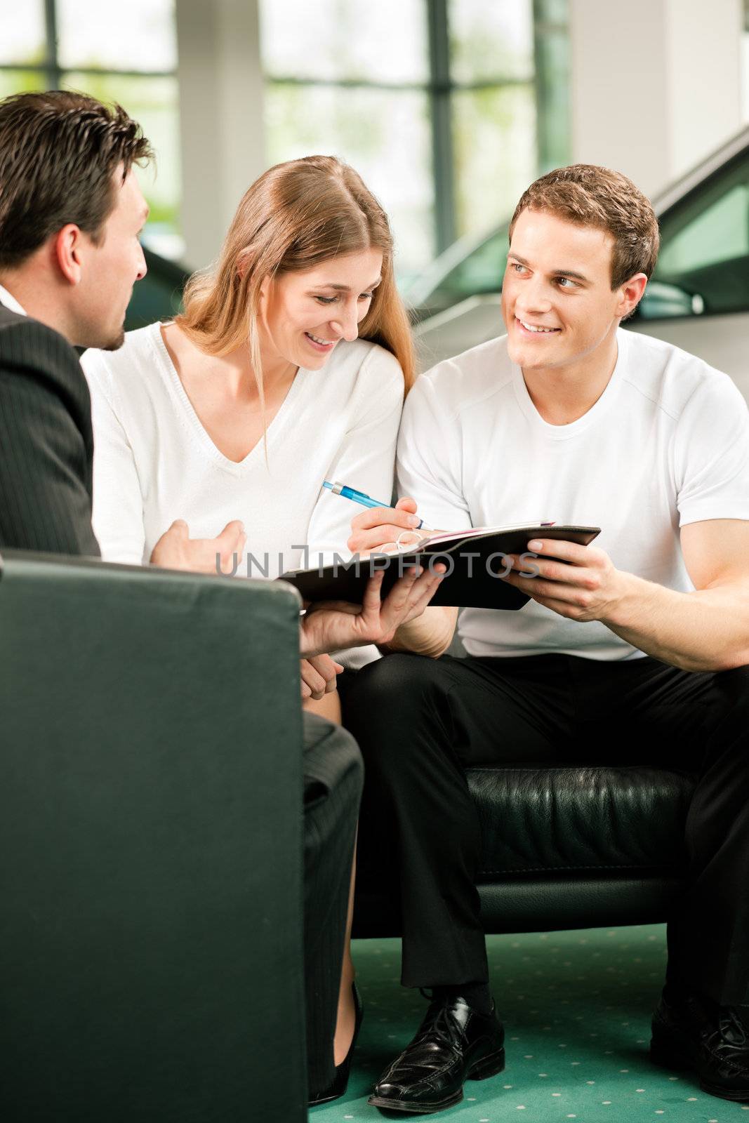Sales situation in a car dealership, the young couple is signing the sales contract to get the new car in the background