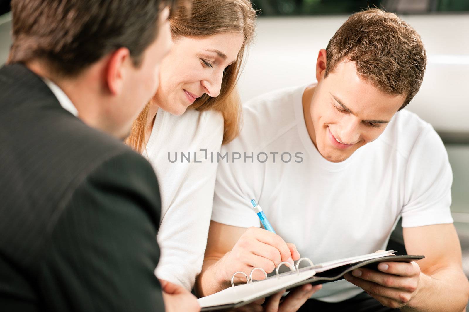 Sales situation in a car dealership, the young couple is signing the sales contract to get the new car in the background