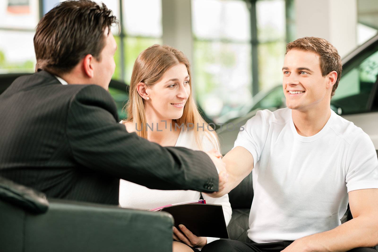 Sales situation in a car dealership, the dealer is shaking hands with a young couple, they want the car in the backgroundSales situation in a car dealership, the dealer is shaking hands with a young couple, they want the car in the background