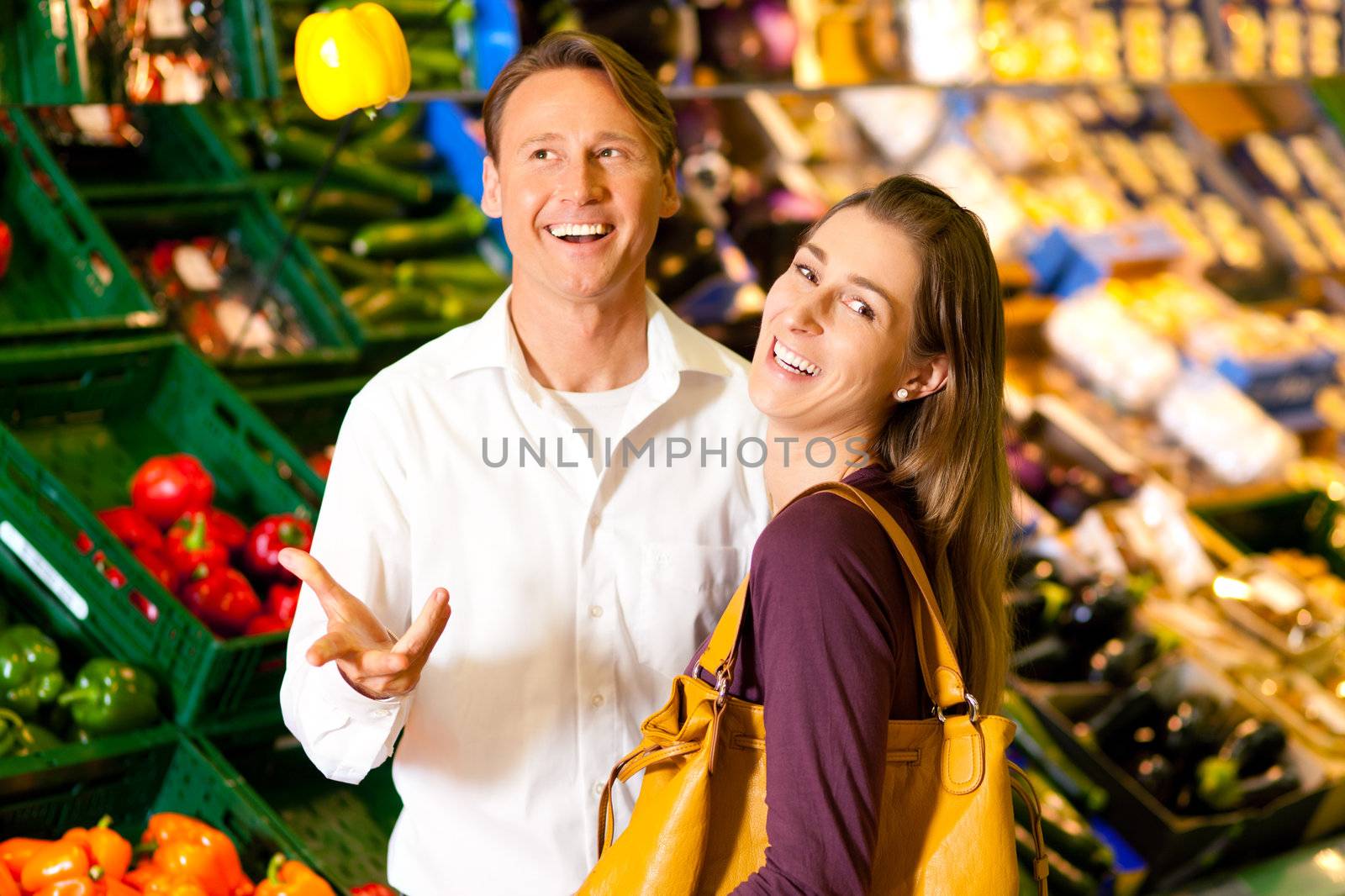 Man and woman in a supermarket at the vegetable shelf shopping for groceries, he is tossing a bell pepper and seems to be full of joy