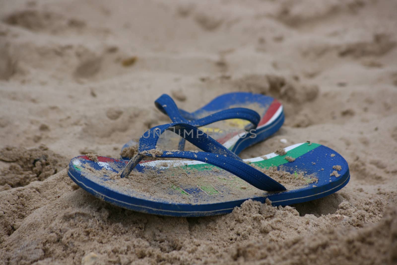 A blue pair of flip flops, or beach sandles, covered in sand and set in the sand on a beach.